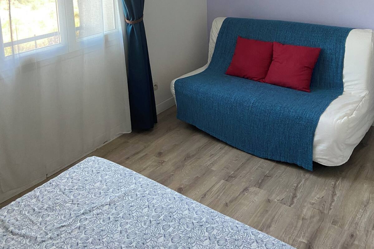A room with a bed, a blue sofa with red cushions, a white shelf, and a window with curtains.