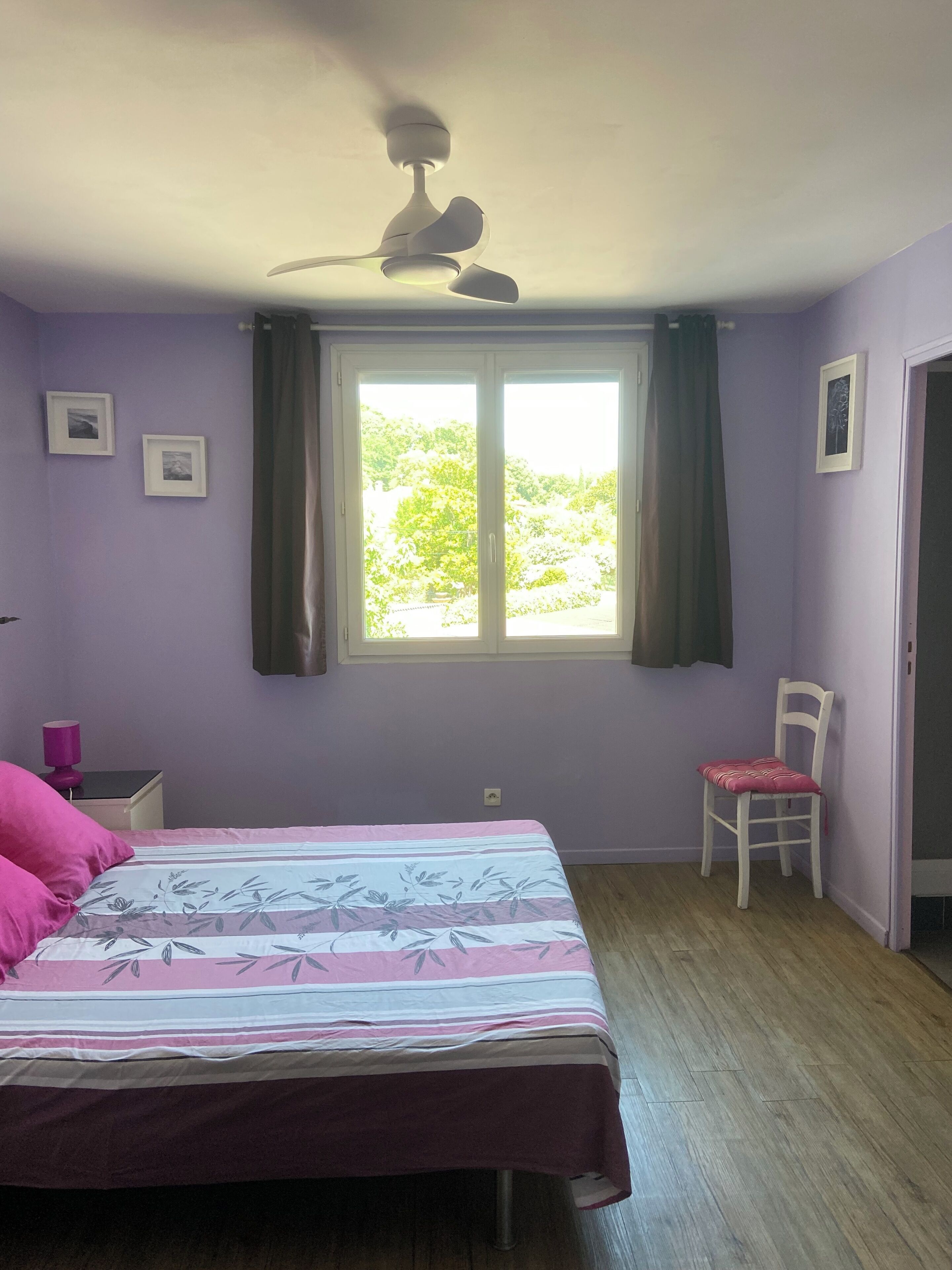 Une chambre à coucher avec un lit, une chaise, un ventilateur de plafond, une fenêtre avec des rideaux et une porte.