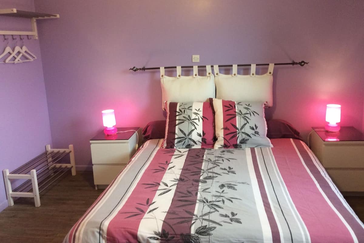 A bedroom with a bed, two bedside tables, a nightstand, a wall-mounted shelf, and a hanging rack.