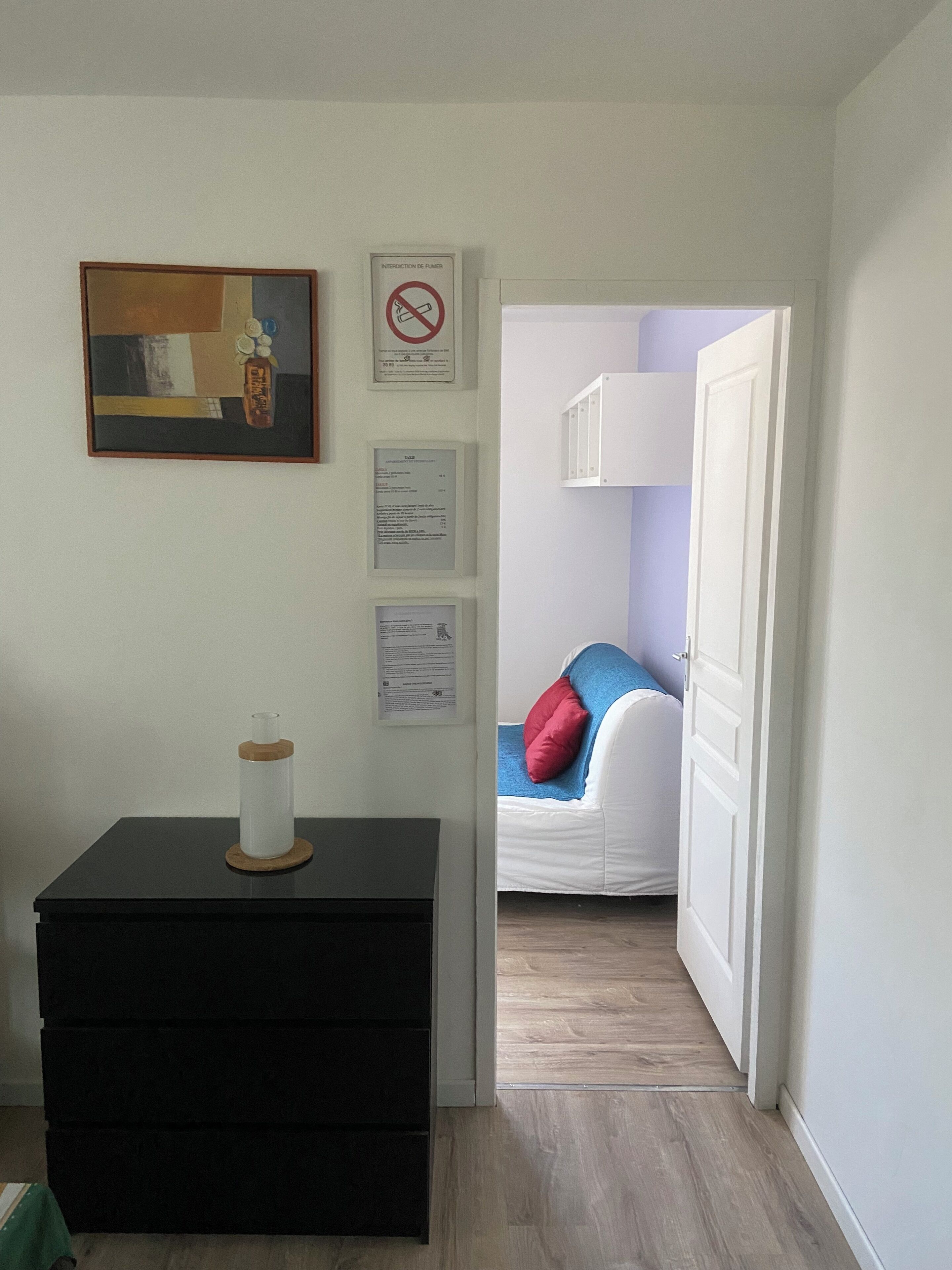Ein Zimmer mit einem schwarzen Kommode, einem Bild, einem Rauchverbotsschild und einem Bett mit blau-roten Kissen.