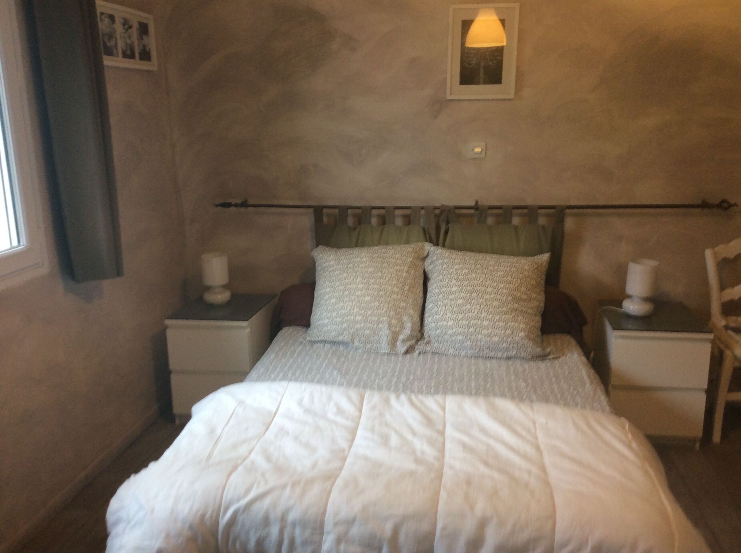 A bedroom with a bed, two bedside tables, a nightstand, a wall-mounted lamp, and a window with curtains.