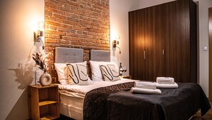 A neatly made bed with a dark quilt, a brick wall, and a wooden wardrobe. - Wine Apartments (Kraków)