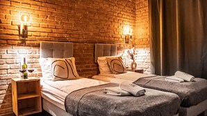 A hotel room with two beds, a brick wall, and a wooden nightstand with a bottle and glass. - Wine Apartments (Kraków)