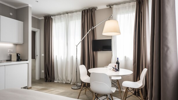 A modern hotel room with a bed, a round dining table with four chairs, a flat-screen TV mounted on the wall, and a floor lamp. - MYSWEETPLACE - Largo Europa Apartments (Padova)