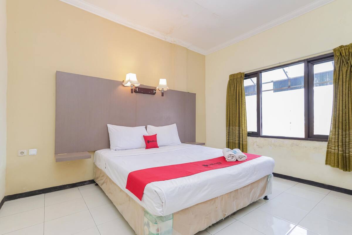 Una habitación de hotel con cama, dos lámparas de pared, un ventanal con cortinas y un cabecero.