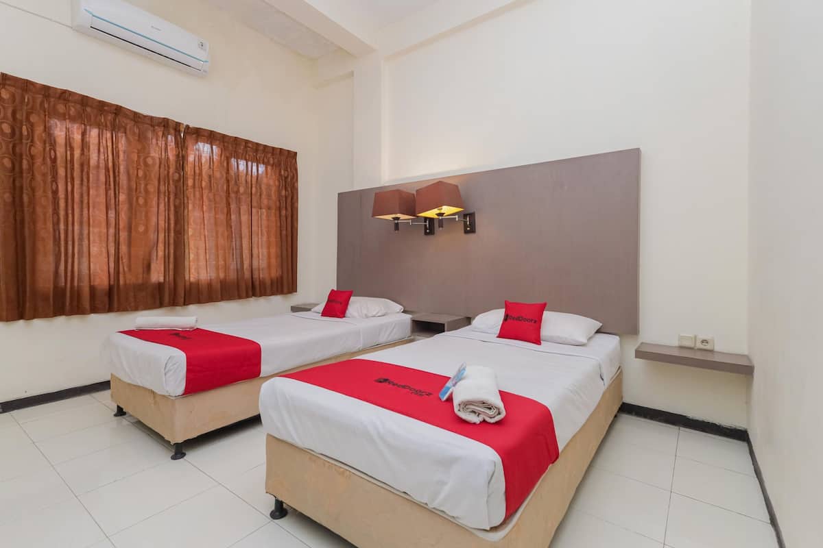 Habitación de hotel con dos camas, cada una con una manta y almohadas rojas, un ventanal con cortinas, una lámpara de pared y un pequeño estante con un reloj.