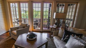 Ein Wohnzimmer mit einer Couch, einem Sessel und einem Tisch mit einer dekorativen Schale. Große Fenster und ein Ausblick ins Freie sind vorhanden.