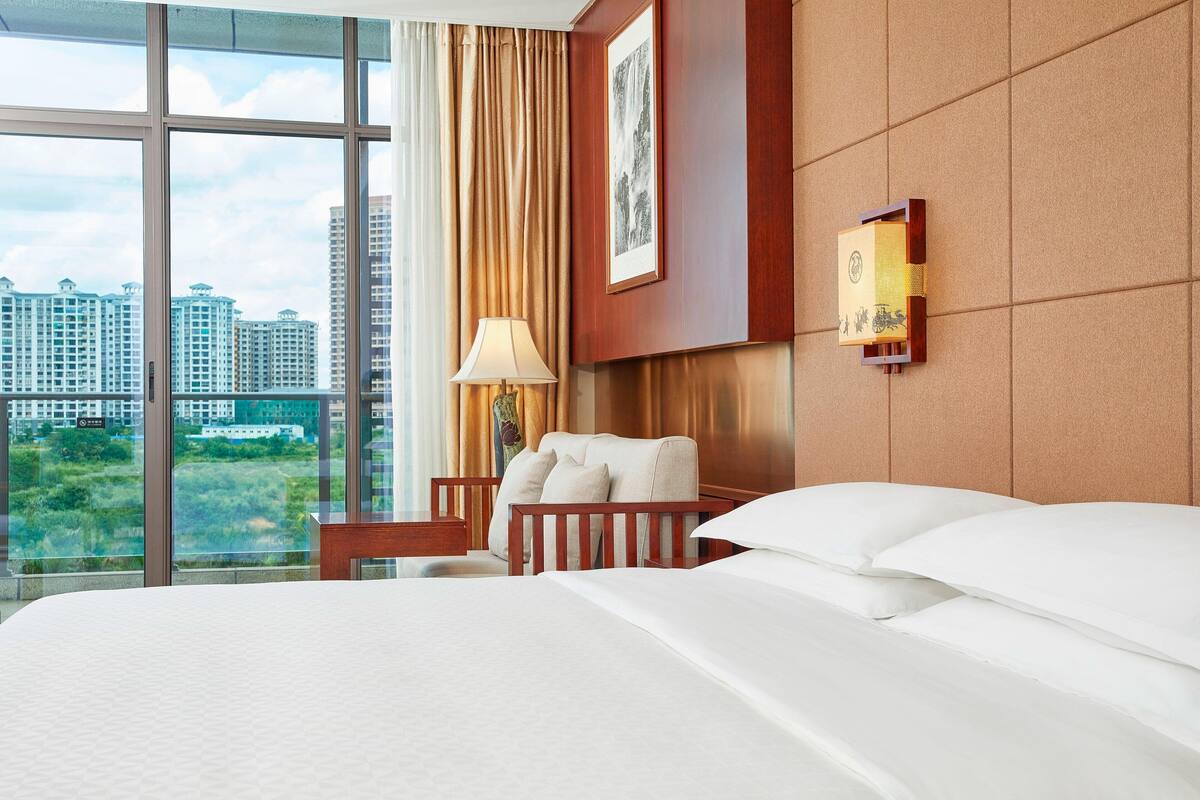 Ein Hotelzimmer mit einem großen Bett, einem Nachttisch, einer Lampe, einem Sessel und Blick auf Gebäude durch große Fenster.