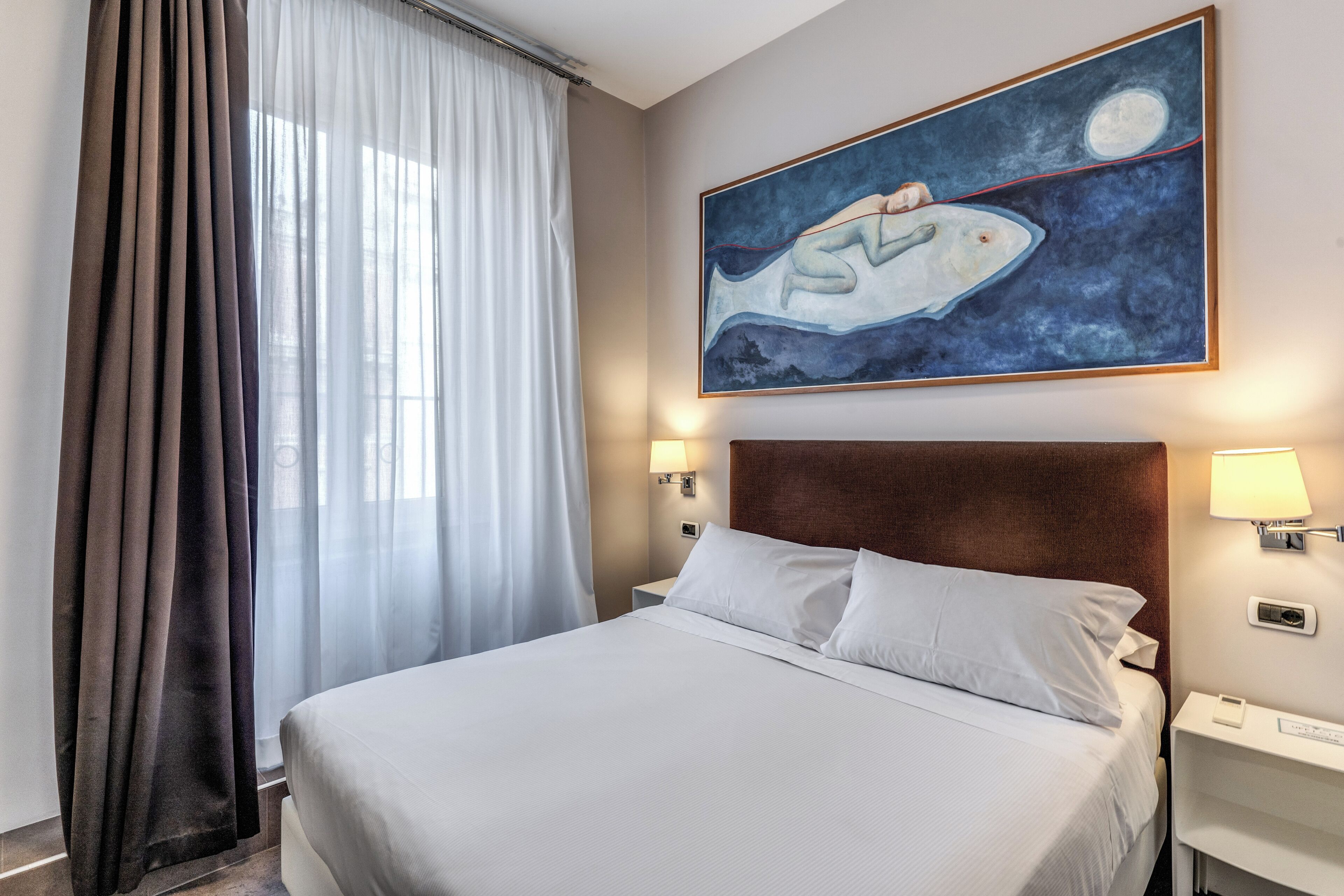 Habitación de hotel con cama, mesita de noche, lámpara, un cuadro y ventana con cortinas.