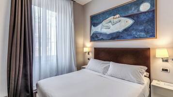 Habitación de hotel con cama, mesita de noche, lámpara, un cuadro y ventana con cortinas.