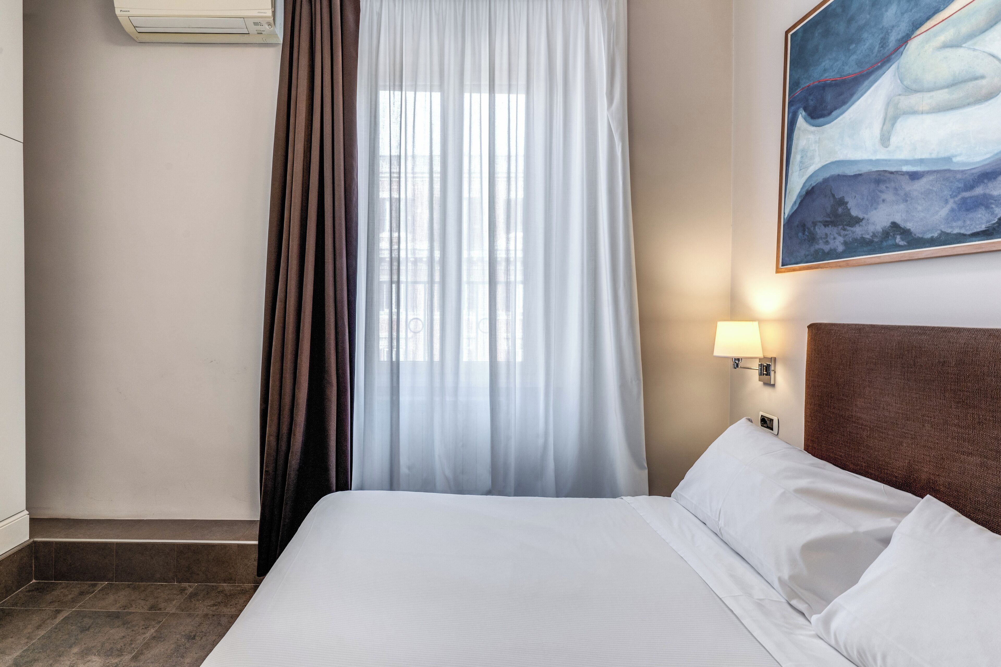 A hotel room with a bed, a window with sheer curtains, a wall-mounted air conditioner, a painting, and a bedside lamp.