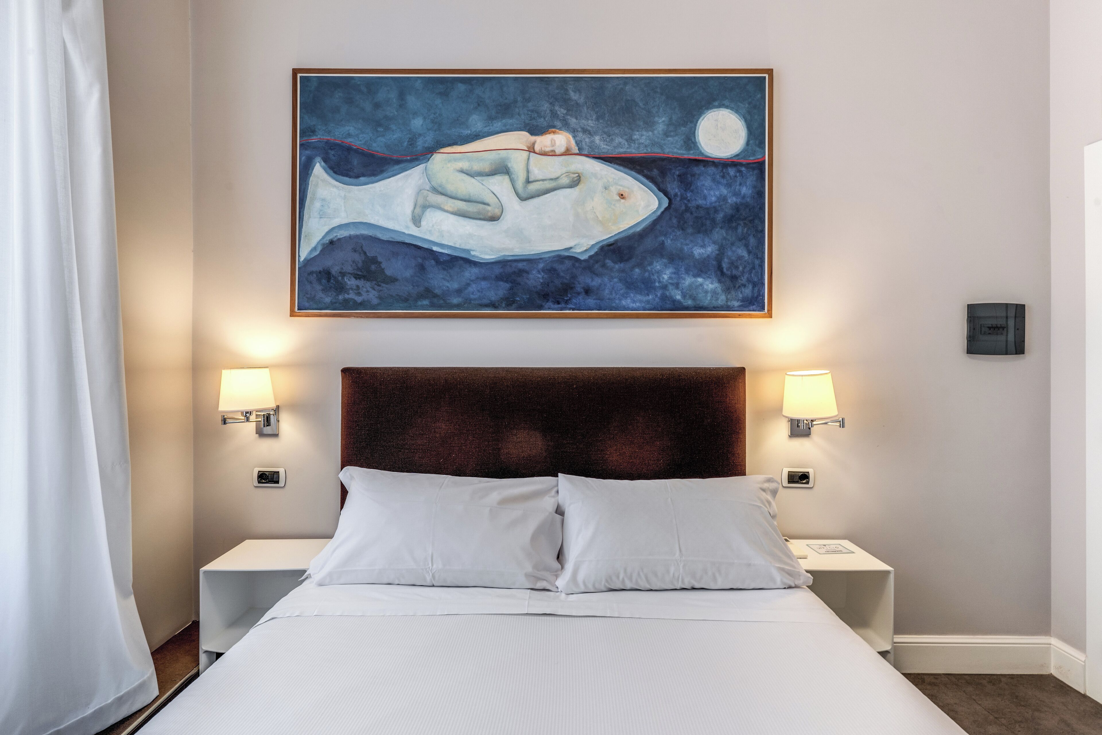 Ein Hotelzimmer mit einem großen Bett, zwei Nachttischen, einem Wandbild und einem Fenster mit Vorhängen.