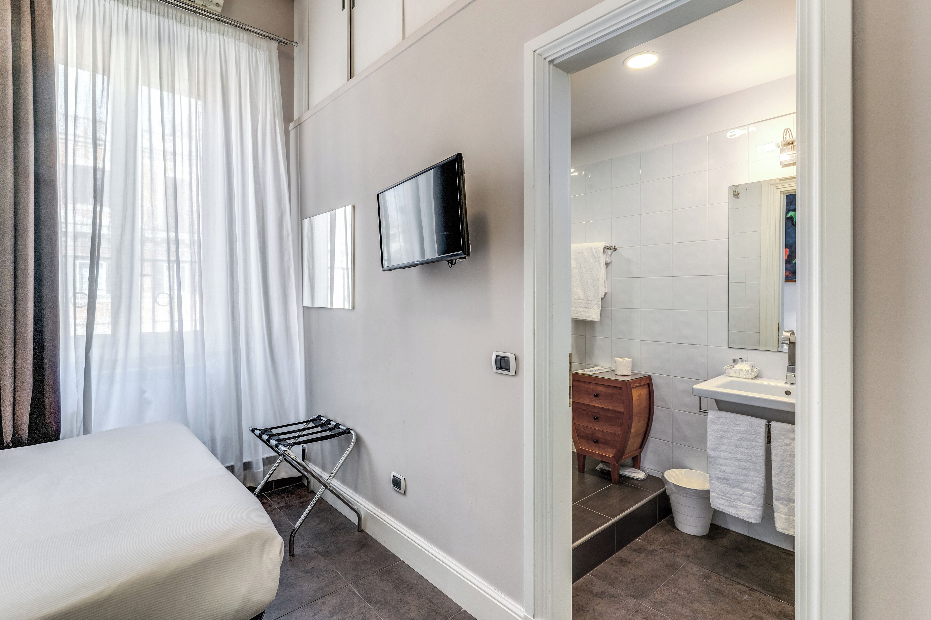 A hotel room with a bed, a TV mounted on the wall, a bathroom with a sink and mirror, and a window with sheer curtains.