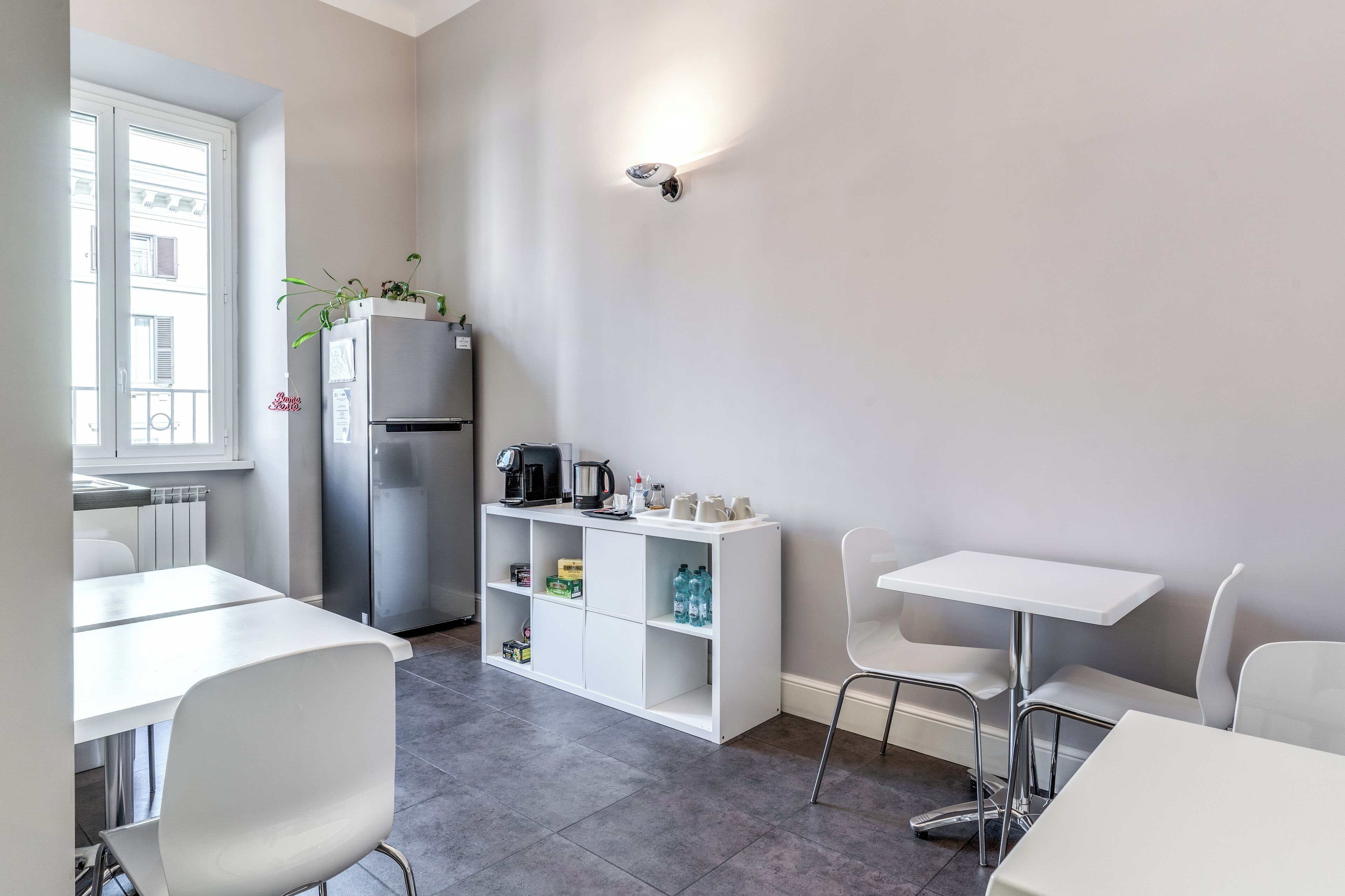 A modern kitchen with white cabinetry, a built-in refrigerator, and a coffee machine. There are white chairs and a table, and a window with a view of buildings.