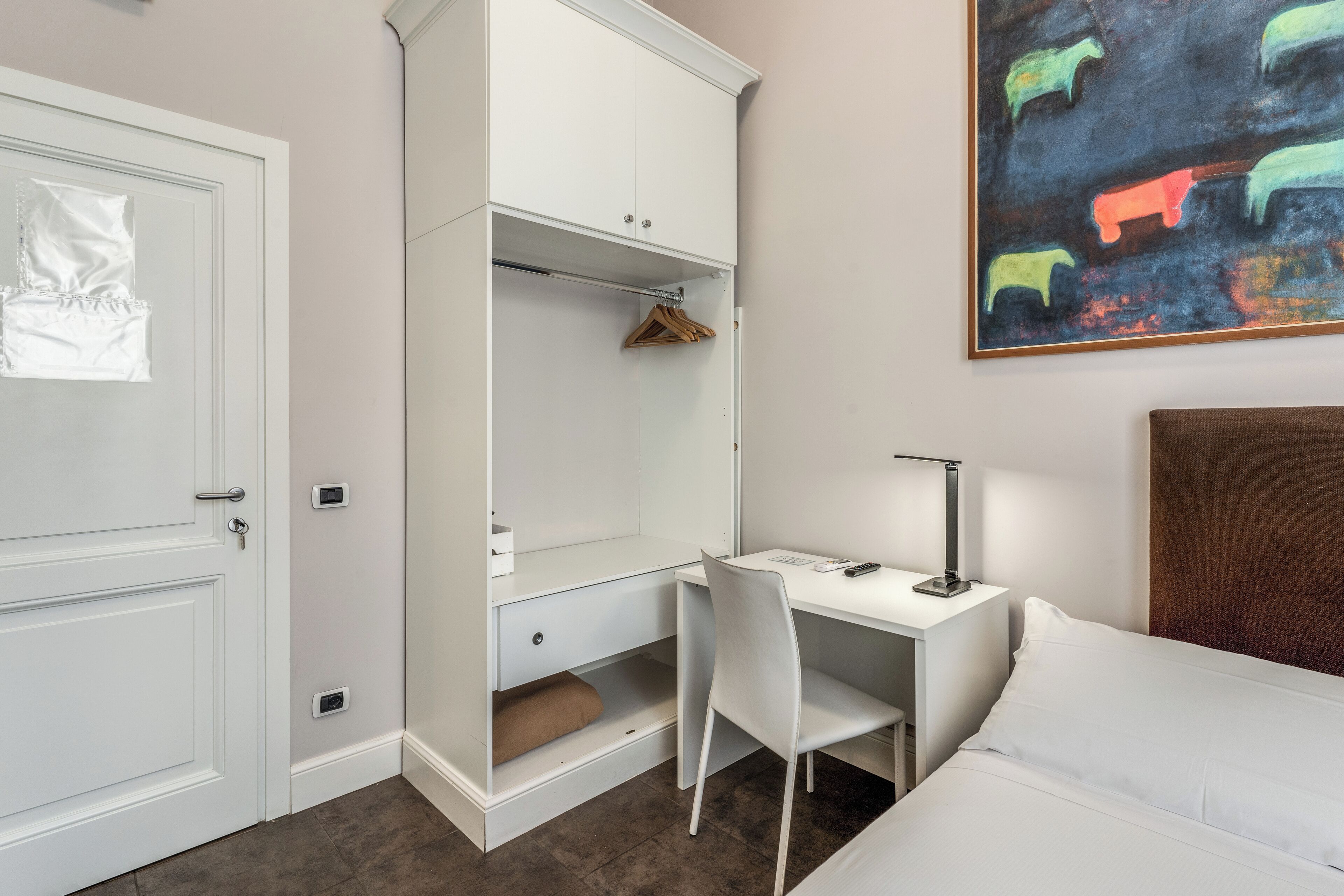 A bedroom with a bed, a desk with a chair, a wardrobe, and a painting on the wall.