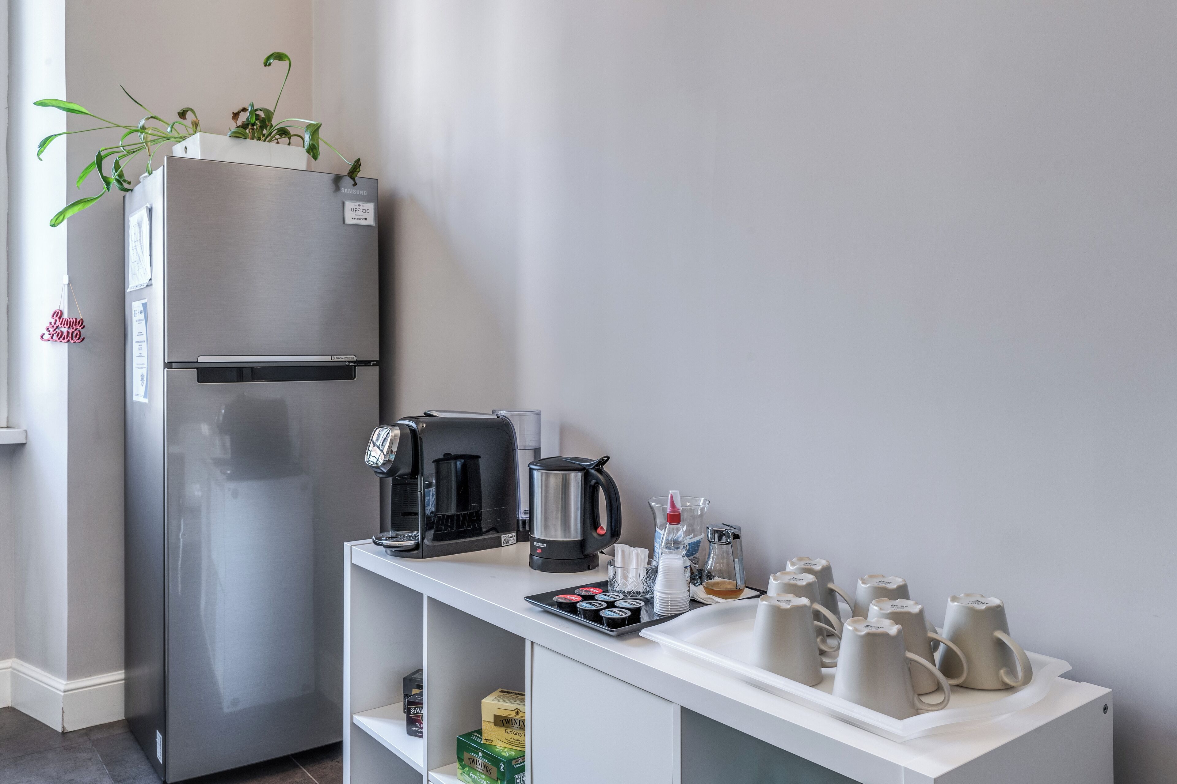 Una kitchenette con un refrigerador de acero inoxidable, una cafetera, un hervidor y varias tazas sobre una encimera blanca.