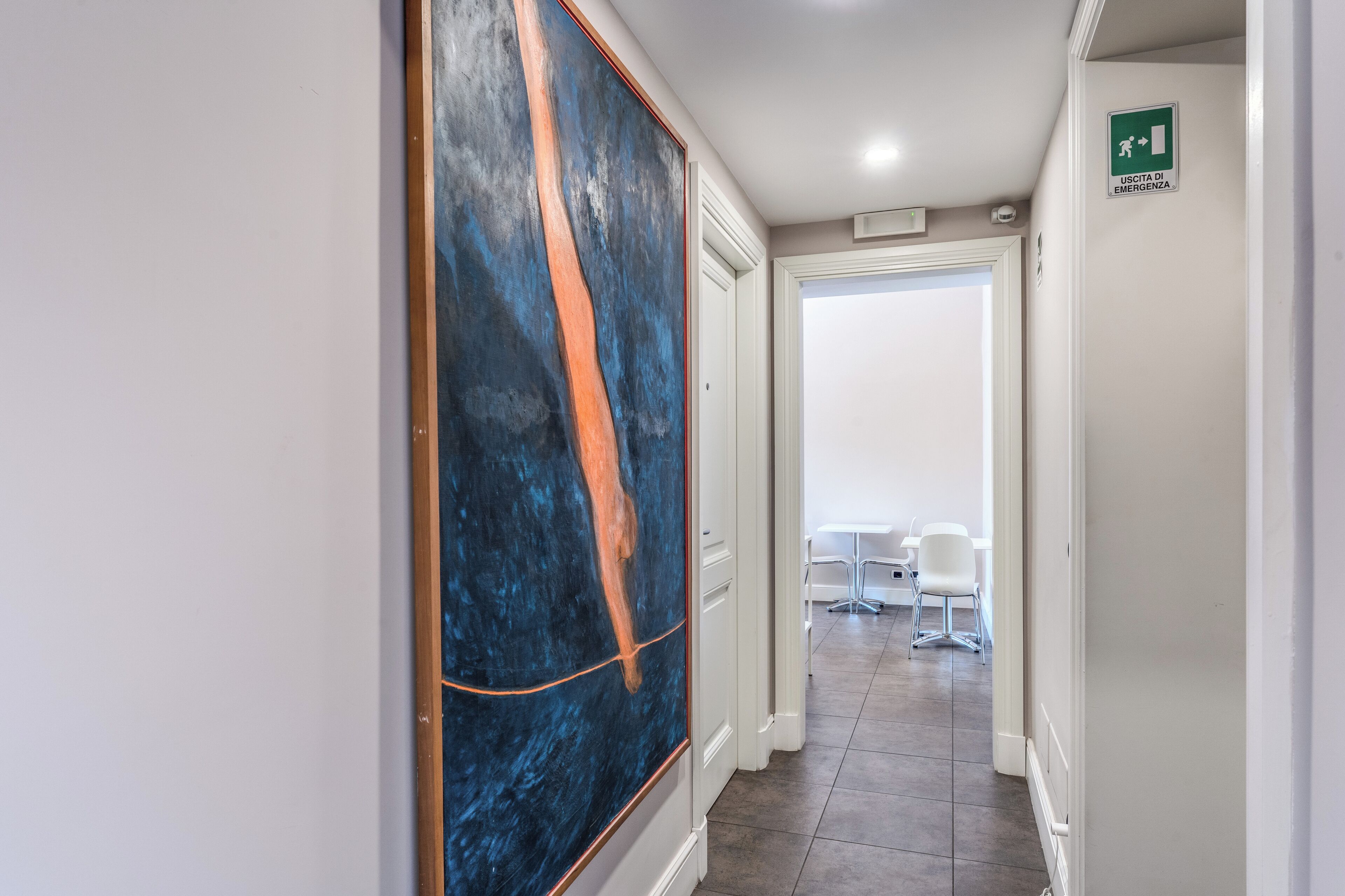 Corredor com uma grande pintura abstrata, uma porta branca e uma placa indicando saída de emergência.