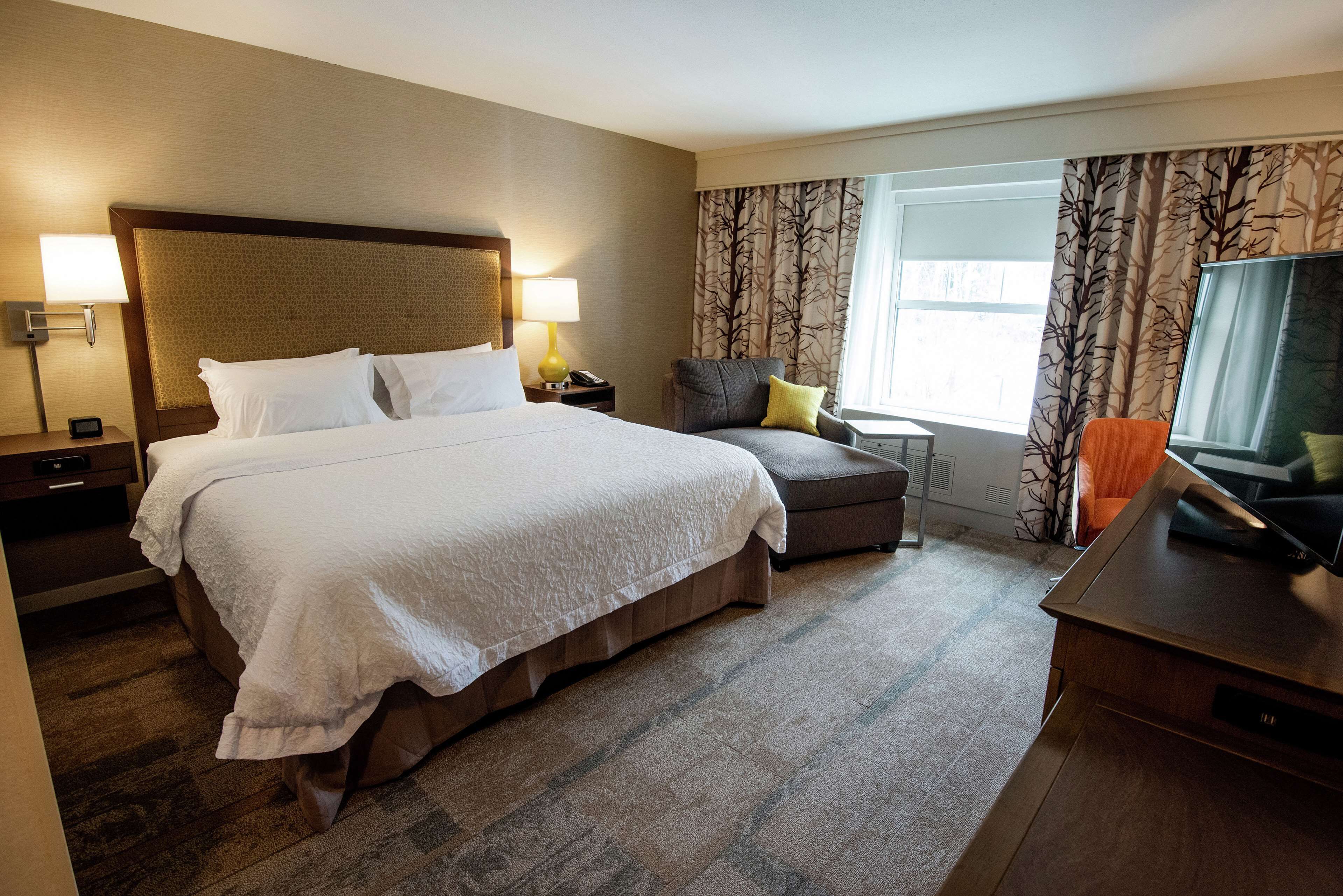 A hotel room with a large bed, a desk, a chair, a TV, and a window with curtains.