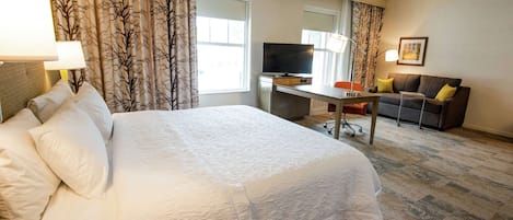 Ein Hotelzimmer mit einem großen Bett, einem Schreibtisch mit Fernseher, einer Couch und einem fensterbepflanzten Fenster.