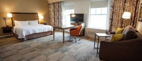 A hotel room with a bed, a desk, a TV, a sofa, and large windows with curtains.