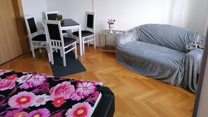 Ein Zimmer mit grauem Sofa, einem weißen Tisch mit Stühlen, einem kleinen Beistelltisch mit Blumen und einer Stehlampe.