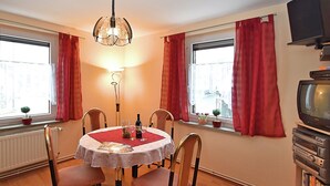 Ein Essbereich mit einem runden Tisch, Stühlen, einer Chandelüster, Fenstern mit roten Vorhängen, einem Heizkörper und einem Fernseher auf einem Schrank.