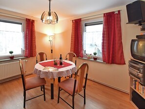 A dining area with a round table, chairs, a chandelier, windows with red curtains, a radiator, and a television on a stand. - Cozy Apartment Near Ski (Muldenhammer)
