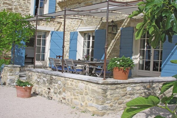 Edificio de piedra con un patio cubierto, que cuenta con una mesa, sillas y plantas en macetas.