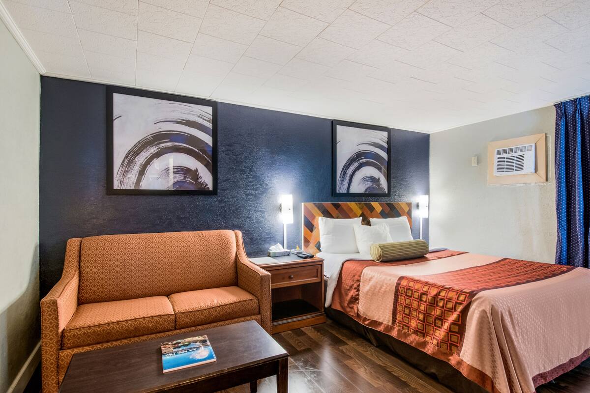 A hotel room with a sofa, a bed with a patterned headboard, a nightstand, and a bedside table.