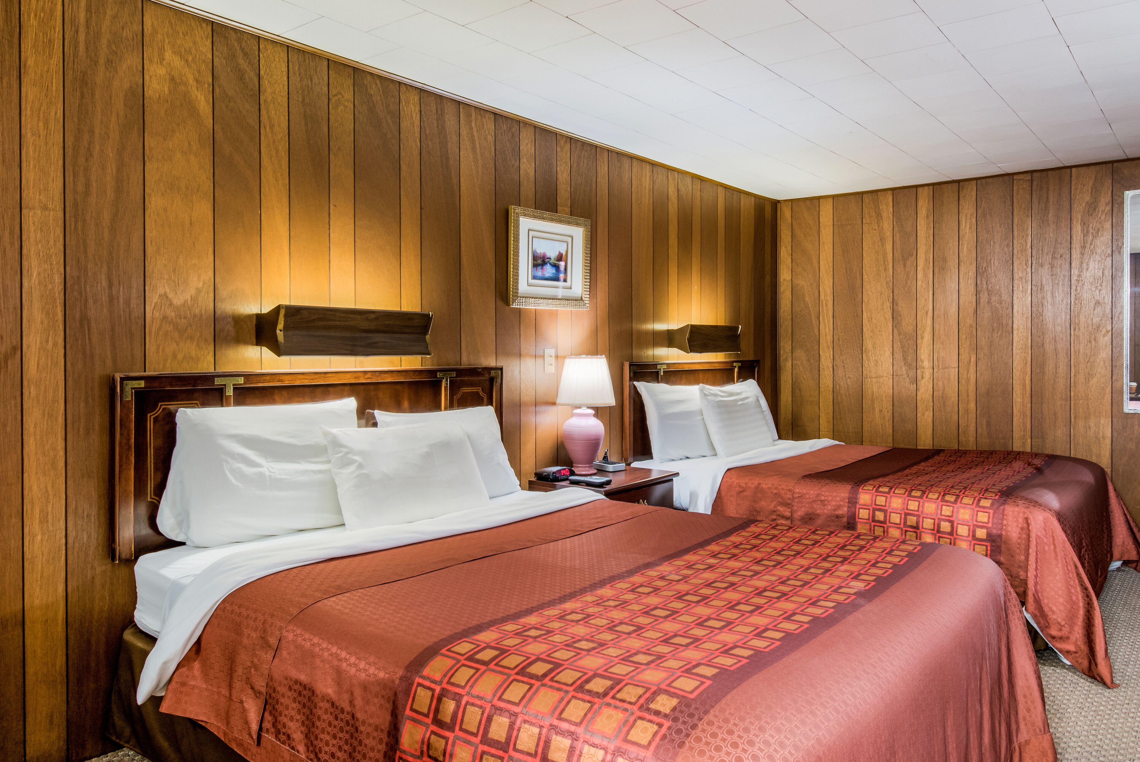 Quarto de hotel com duas camas, paredes com painéis de madeira, um quadro na parede e uma luminária na mesa de cabeceira.