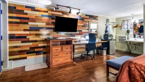 Una habitación con piso de madera, un escritorio con una silla, un televisor montado en una pared de madera multicolores, una cama con una colcha y un tapete pequeño.