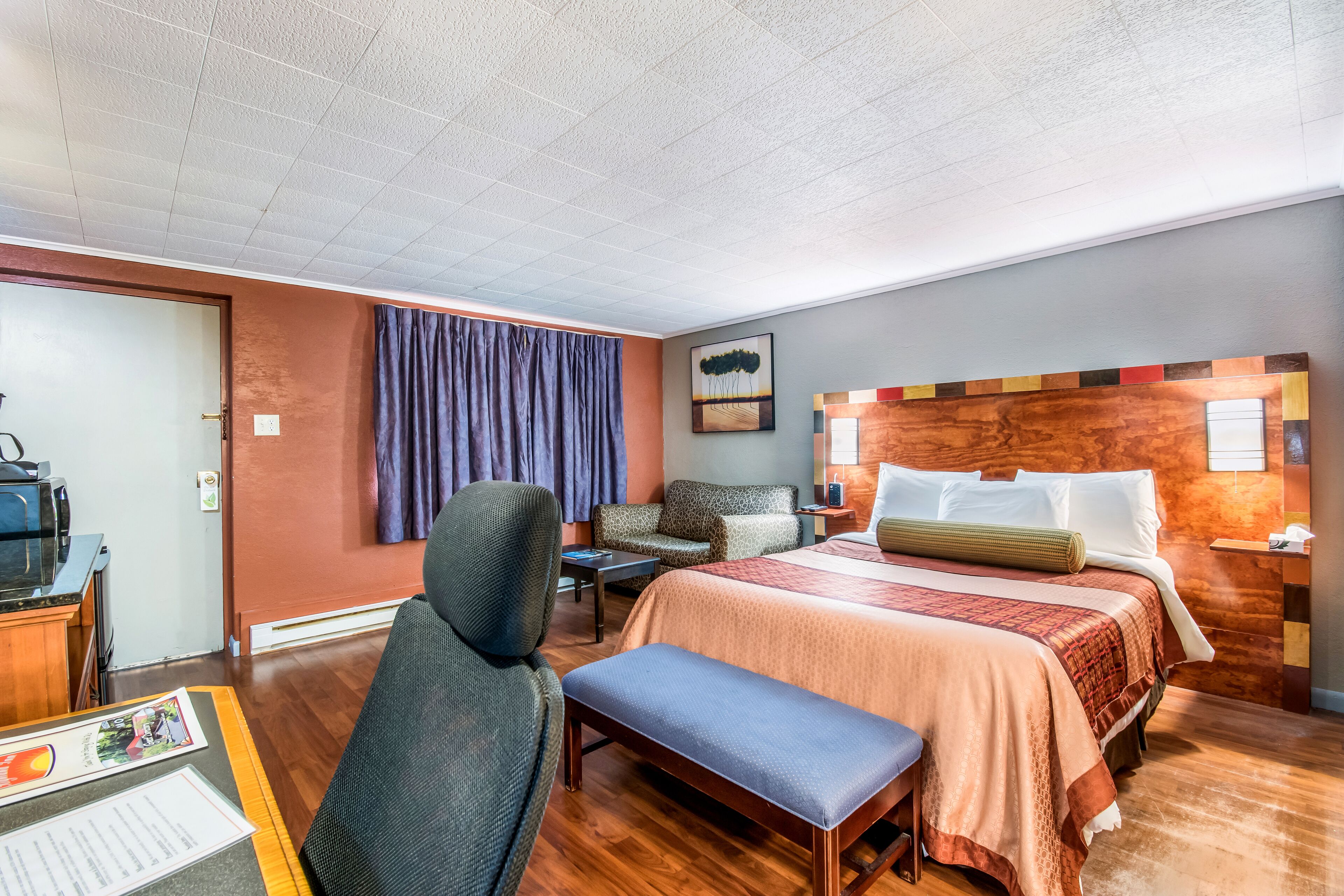 Ein Hotelzimmer mit einem Bett, einem Schreibtisch mit Computer, einem Sessel, einer Bank, einem Fenster mit Vorhängen und einem Wandbild.