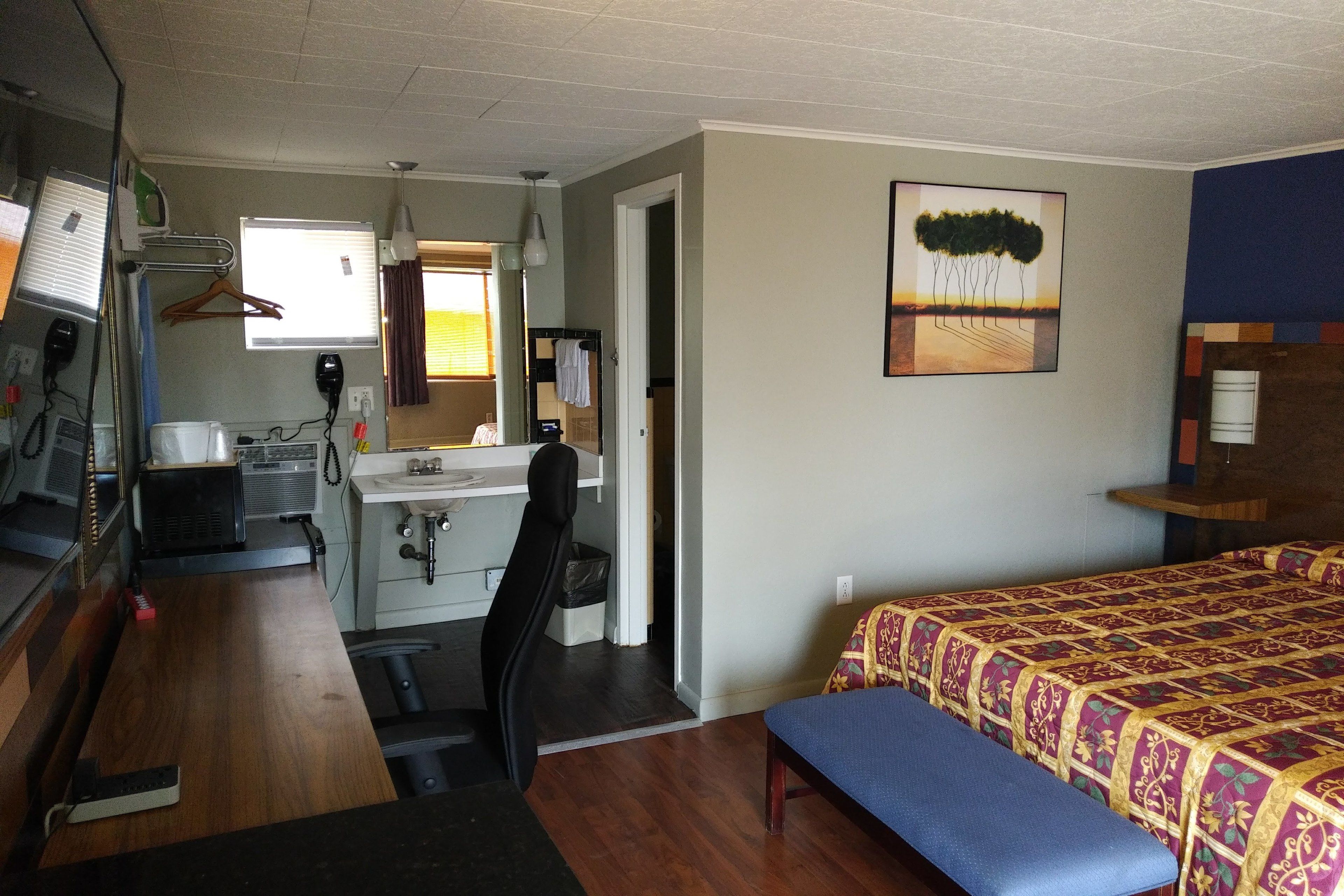 A compact hotel room with a kitchenette, a bed with a patterned bedspread, a bench, a desk with a chair, and a painting on the wall.