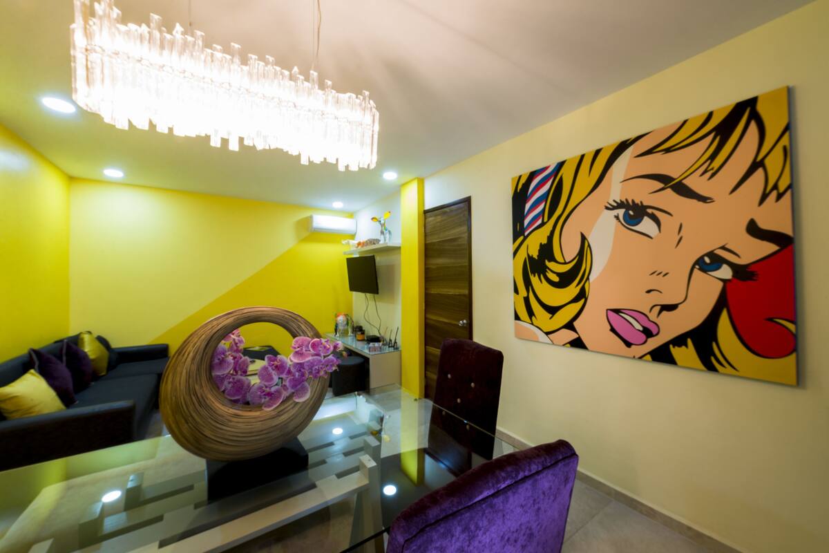 A modern living room with a glass coffee table, a vibrant yellow wall, and a large pop art painting.