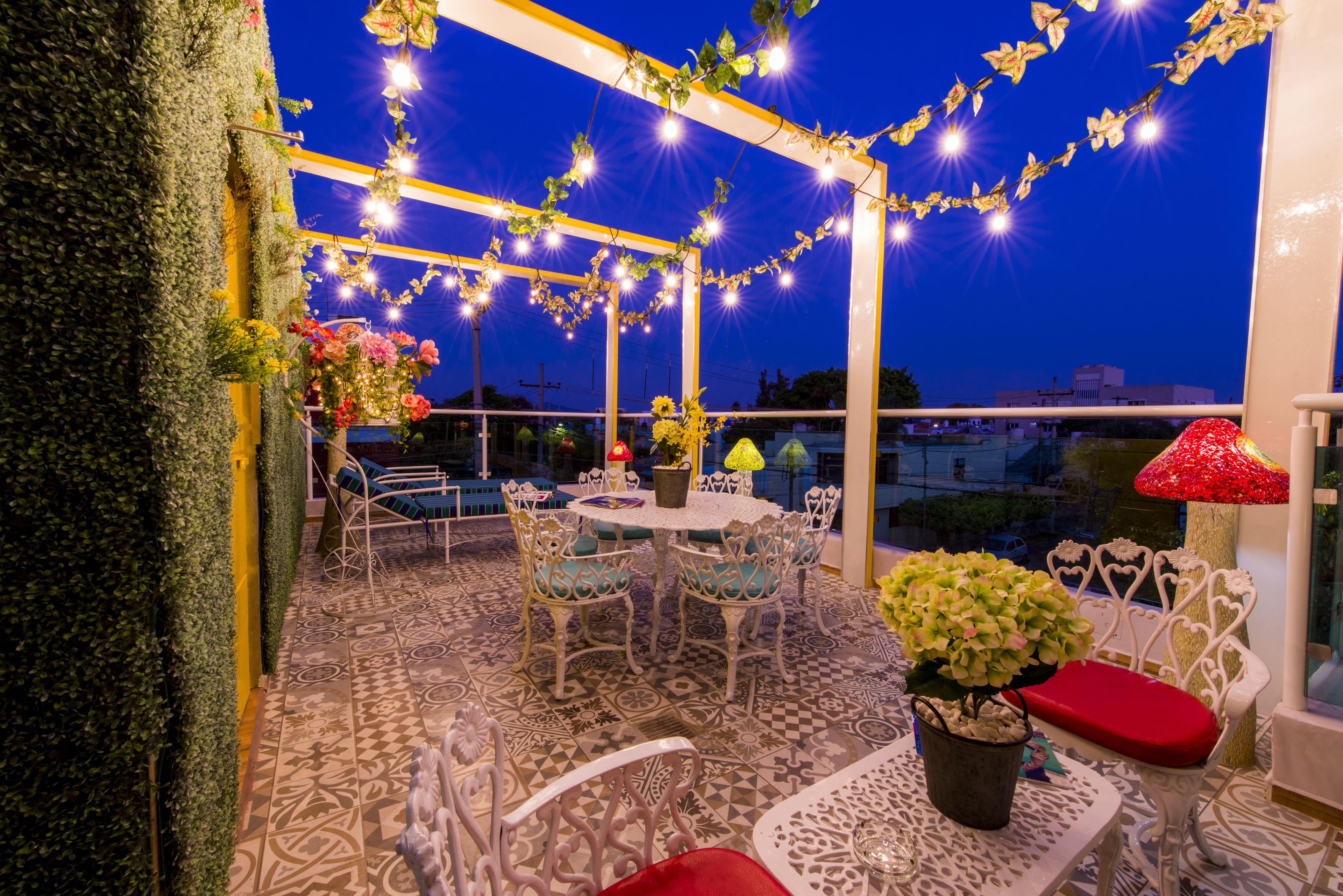 Eine Terrasse im Freien mit einem gemusterten Boden, weißen Metallstühlen, einem runden Tisch und Lichterketten.