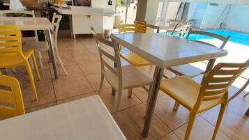 Espaço de refeições com vista para a piscina, incluindo cozinha com relógio e cadeiras amarelas ao redor de uma mesa.