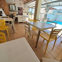 Espaço de refeições com vista para a piscina, incluindo cozinha com relógio e cadeiras amarelas ao redor de uma mesa.