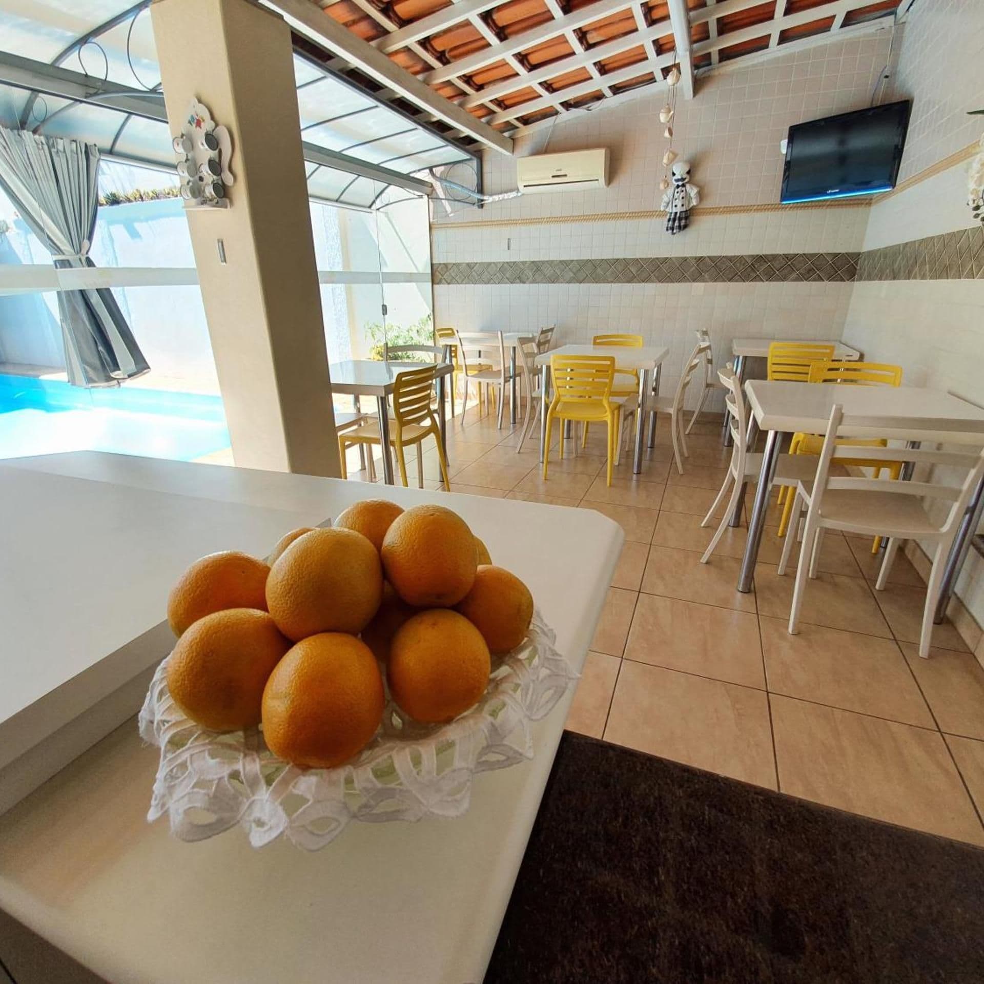 Uma área de refeições com mesas e cadeiras, piscina visível através de um painel de vidro e uma tigela de laranjas sobre uma mesa.