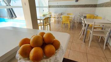 Uma área de refeições com mesas e cadeiras, piscina visível através de um painel de vidro e uma tigela de laranjas sobre uma mesa.