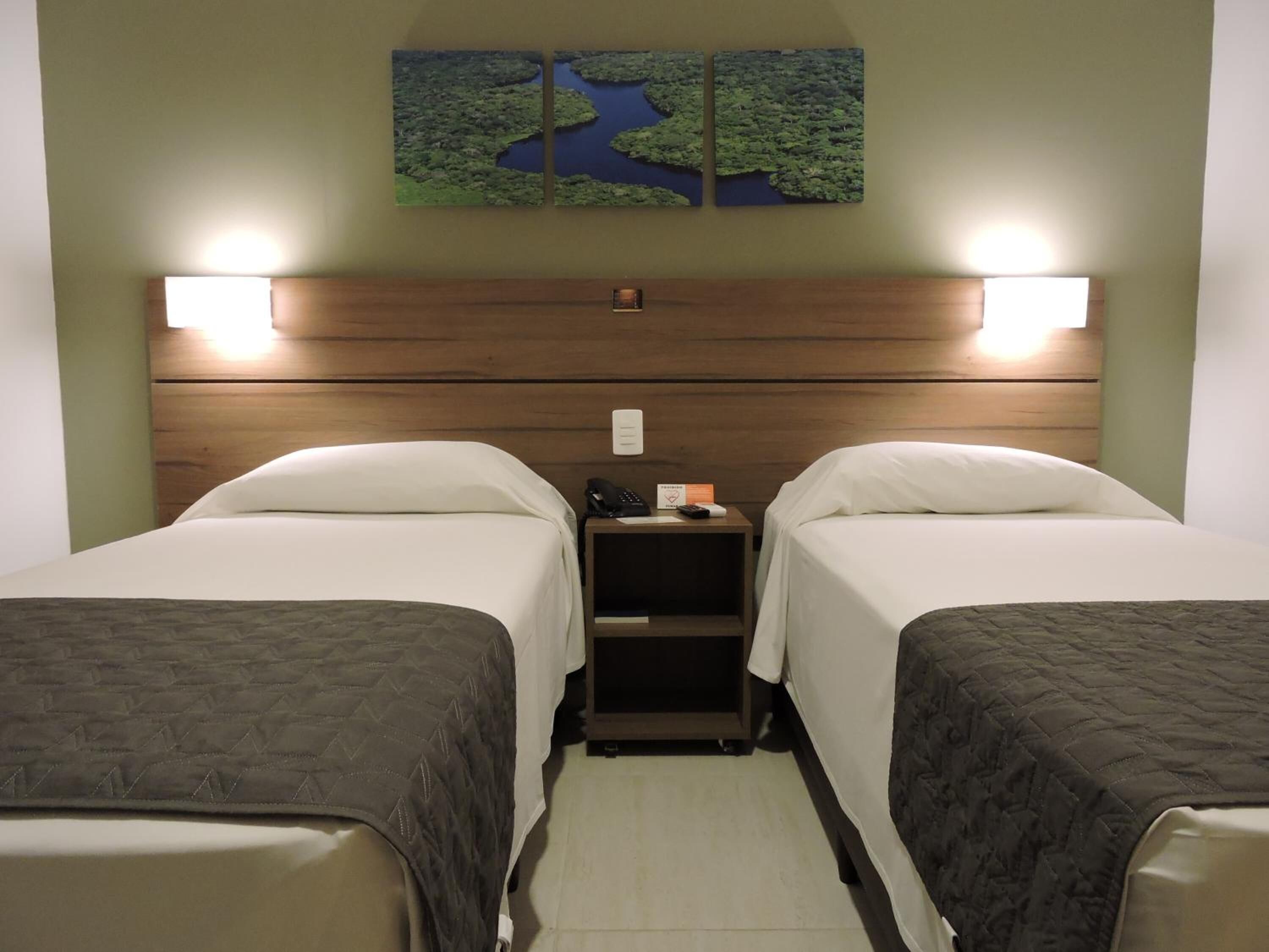 Een hotelkamer met twee bedden, een houten hoofdbord, bedlampjes, een nachtkastje met een telefoon en een bord, en een kunstwerk aan de muur van een landschap.