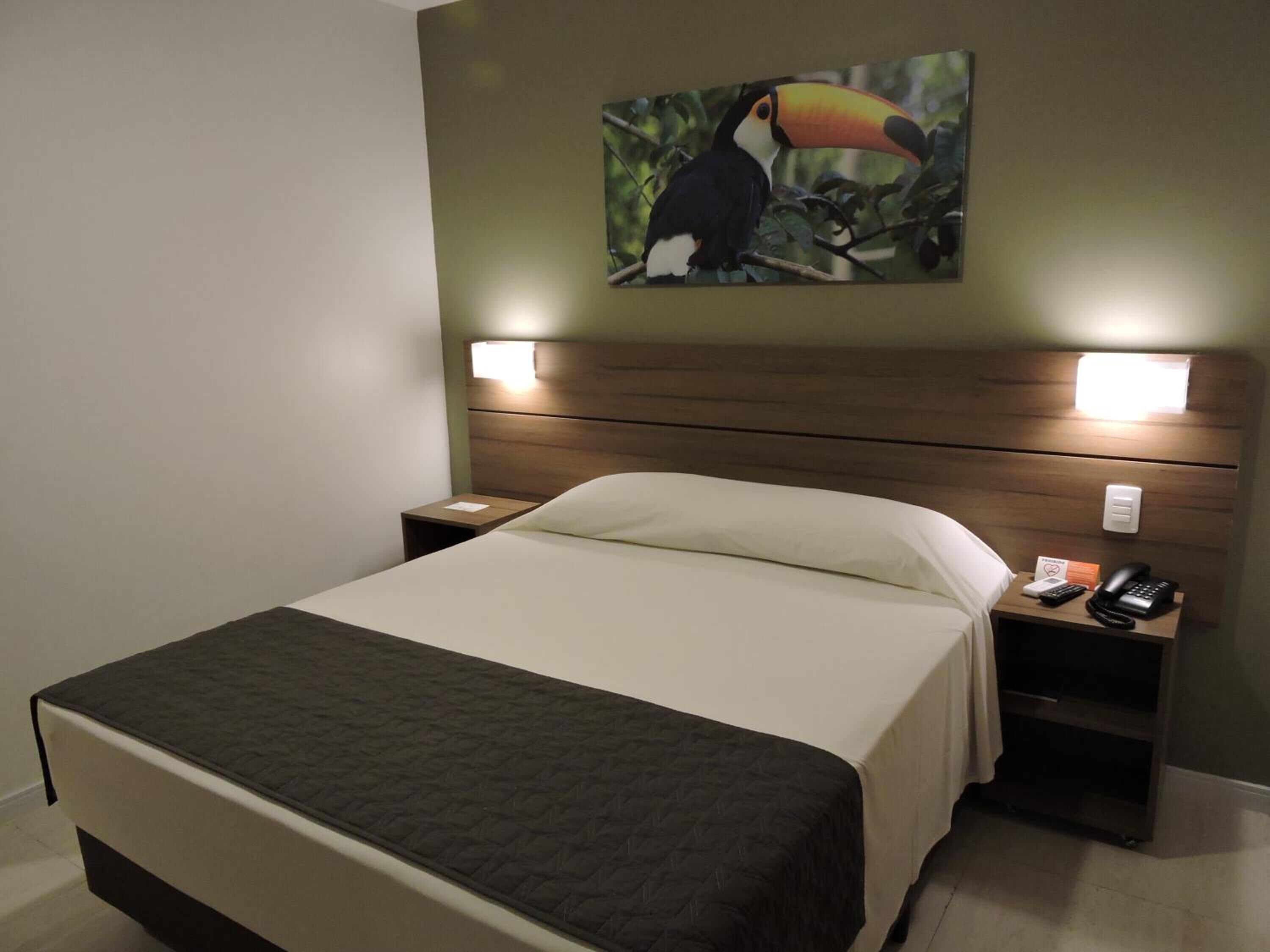 A hotel room with a bed, two bedside tables, a nightstand, a wall-mounted telephone, and a painting of a toucan.