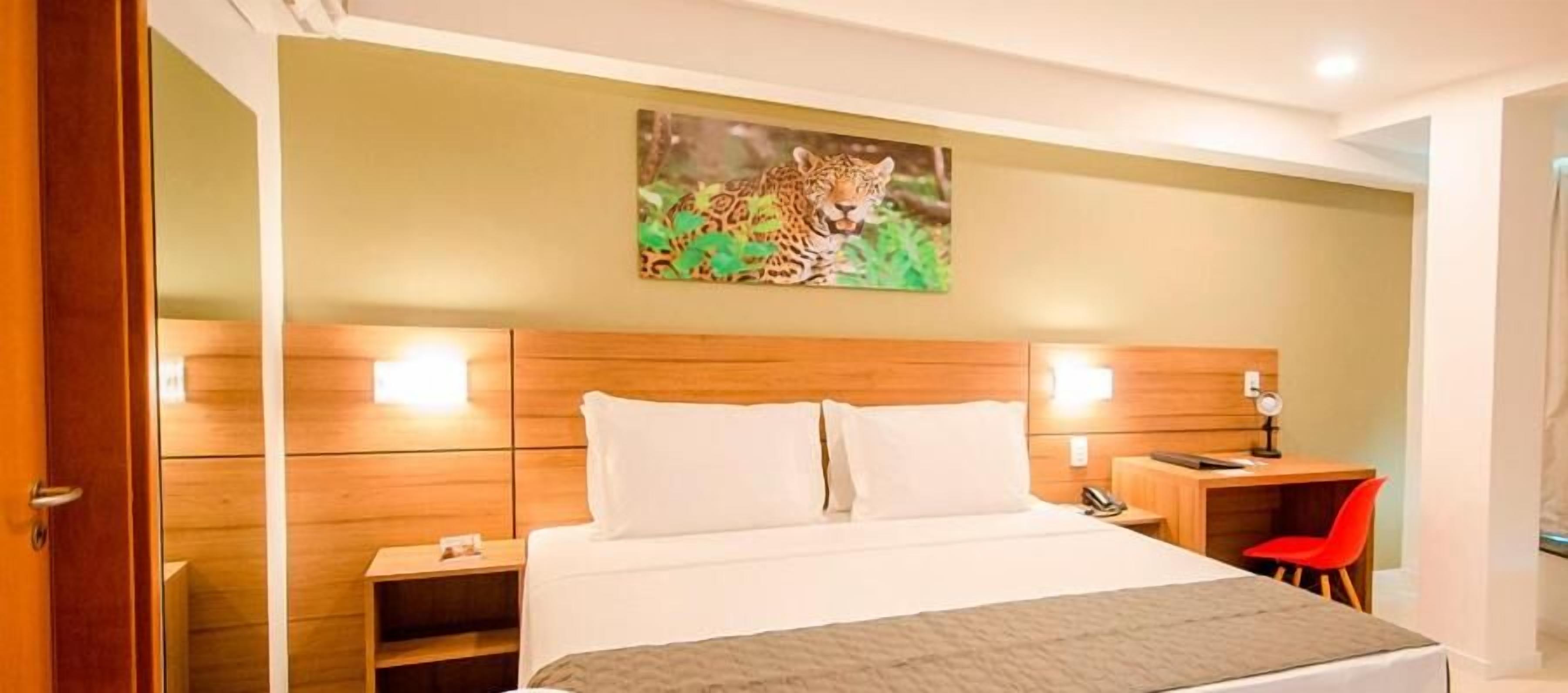 Habitación de hotel con cabecera de madera, cama con ropa blanca, escritorio con silla y un cuadro de un leopardo en la pared.