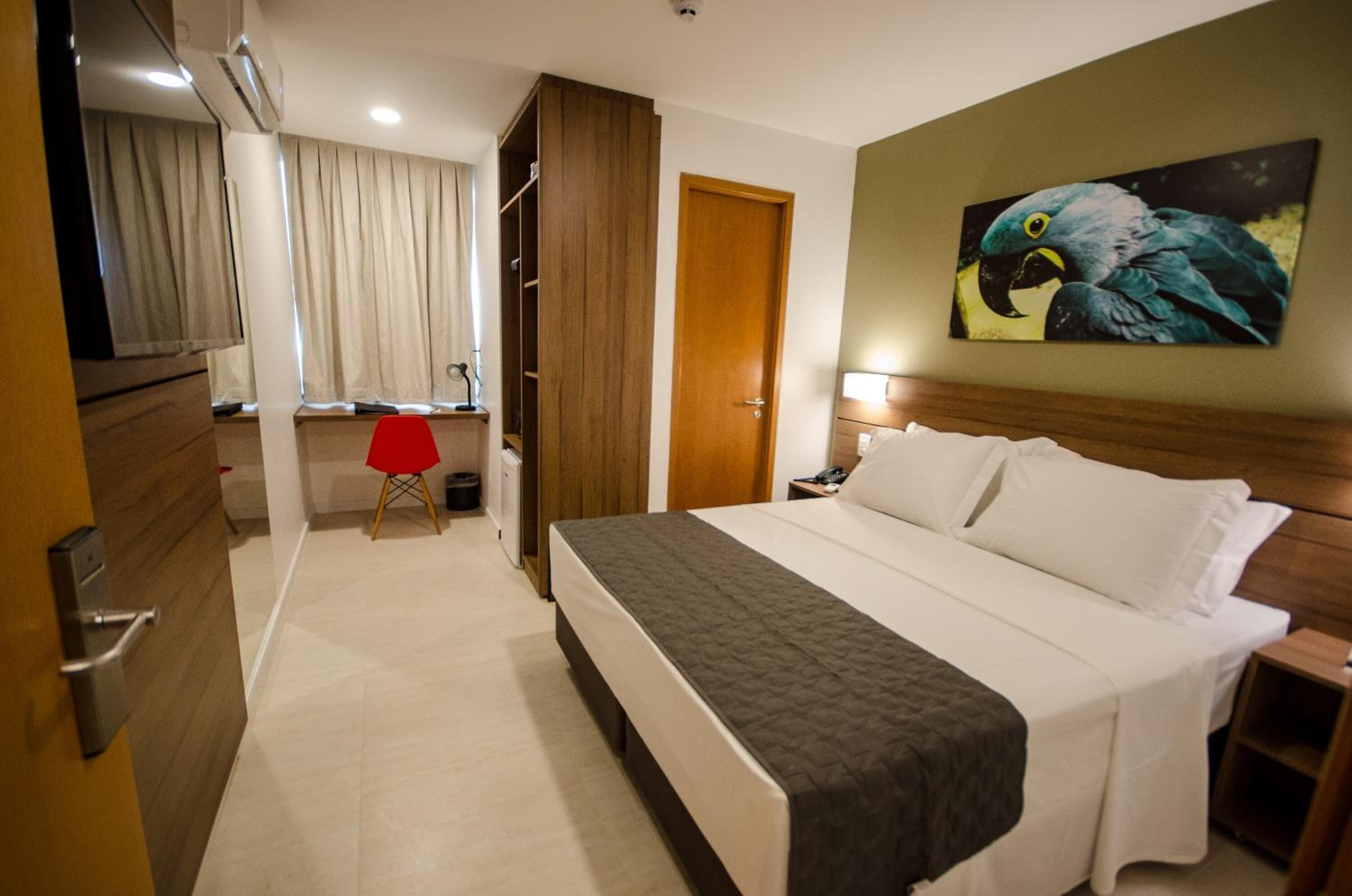 Quarto de hotel com uma cama grande, um guarda-roupa de madeira, uma escrivaninha com cadeira e um quadro de um papagaio na parede.