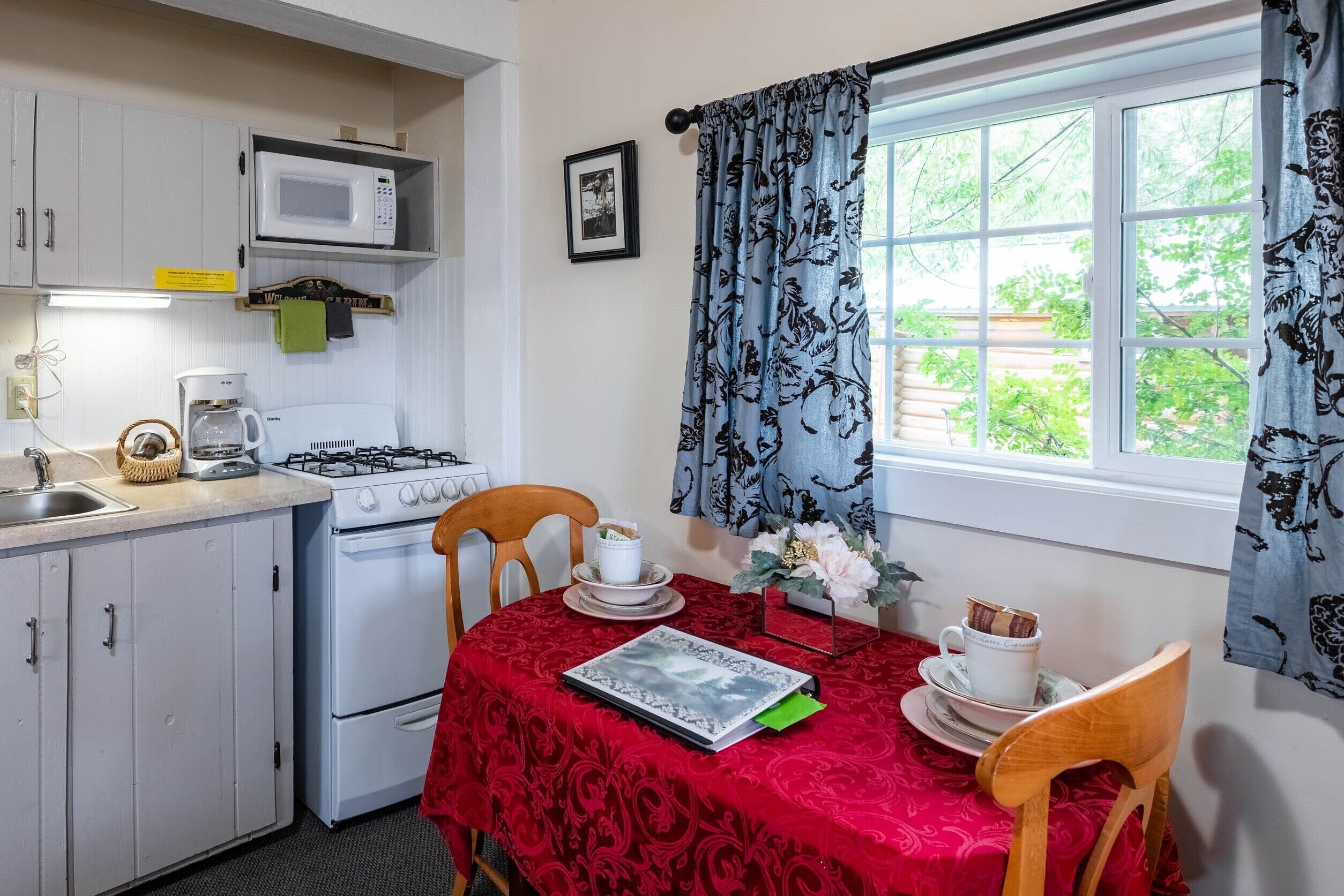 A kitchen with white cabinets, a microwave, a coffee maker, and a stove. There's a dining table with a red tablecloth, a chair, and a cup of coffee.