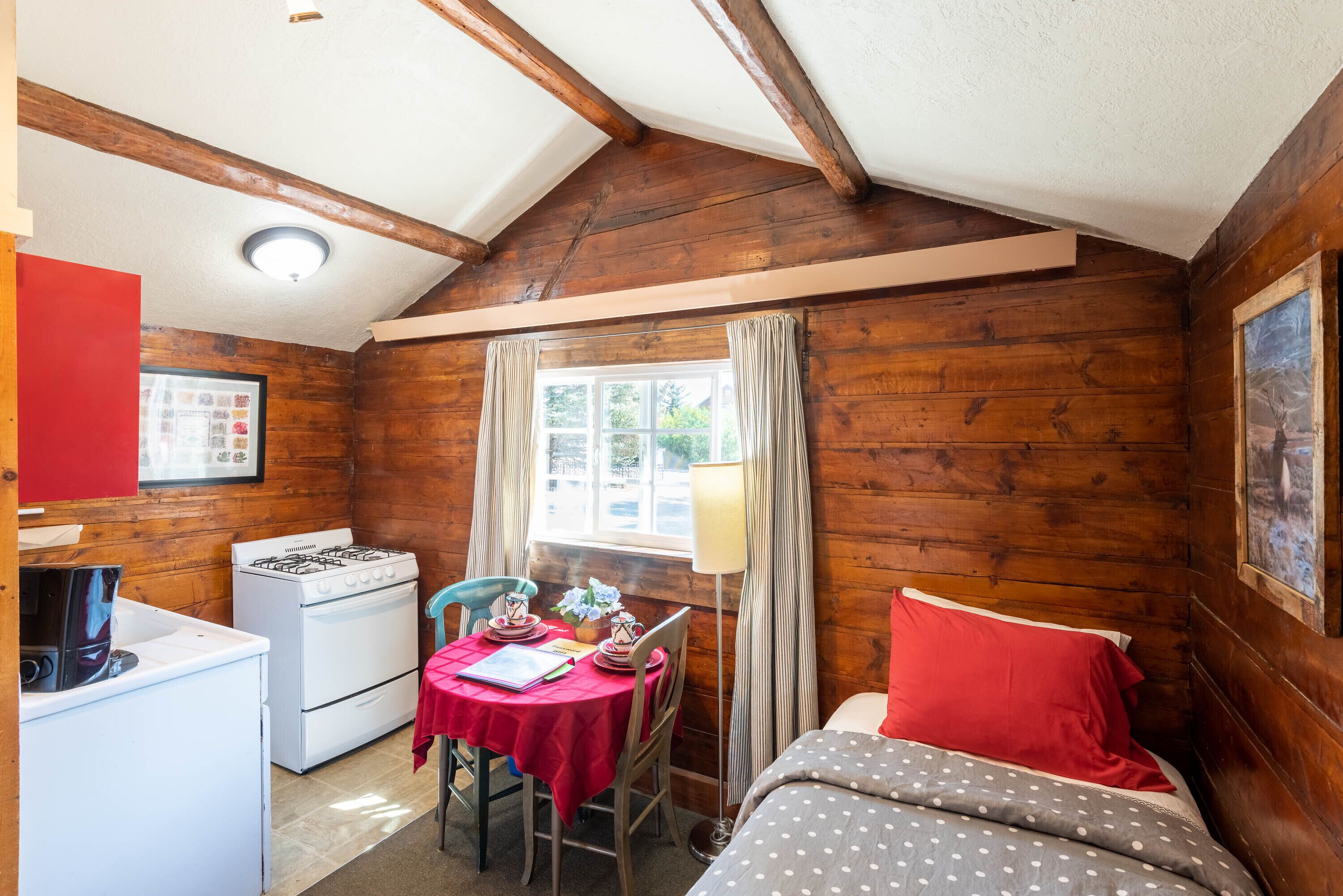 A cozy cabin-style room with a wooden interior, a dining table with chairs, a bed with red pillows, and a window with curtains.