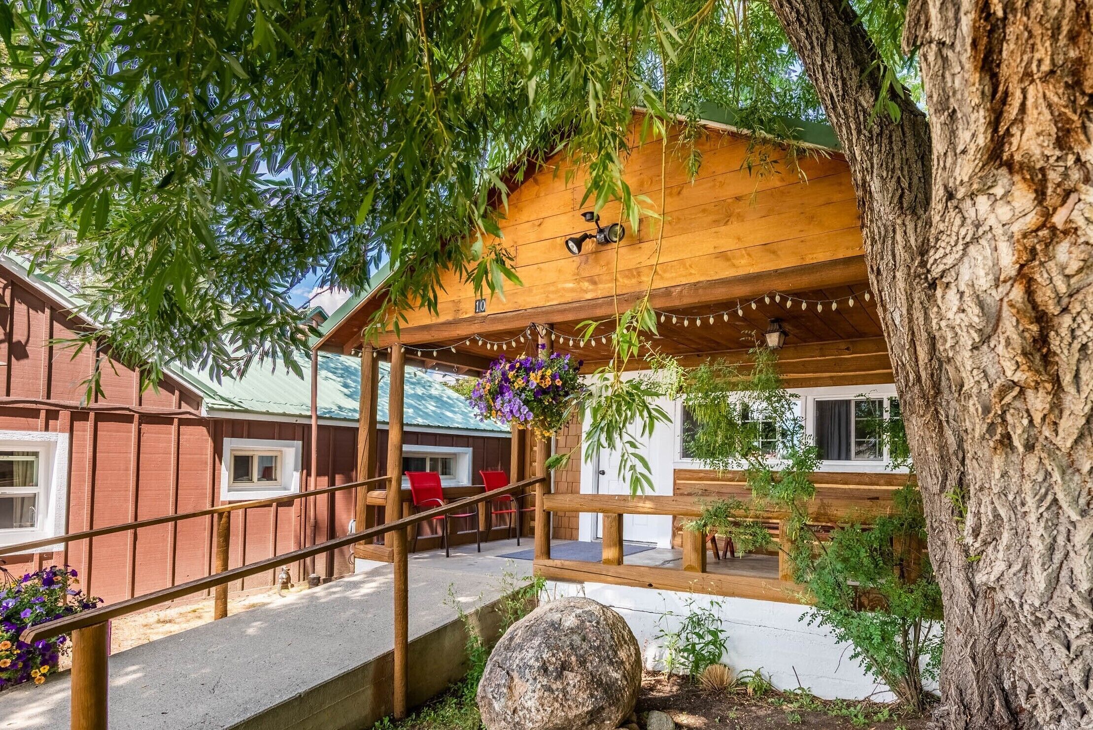 Una cabaña de madera con porche cubierto, mobiliario exterior y un árbol grande al frente.
