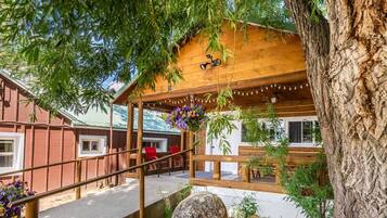 Una cabaña de madera con porche cubierto, mobiliario exterior y un árbol grande al frente.