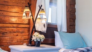 A wooden cabin-style room with a bed, a nightstand with a lamp, and a window with curtains. - Log Cabin Motel (Pinedale)