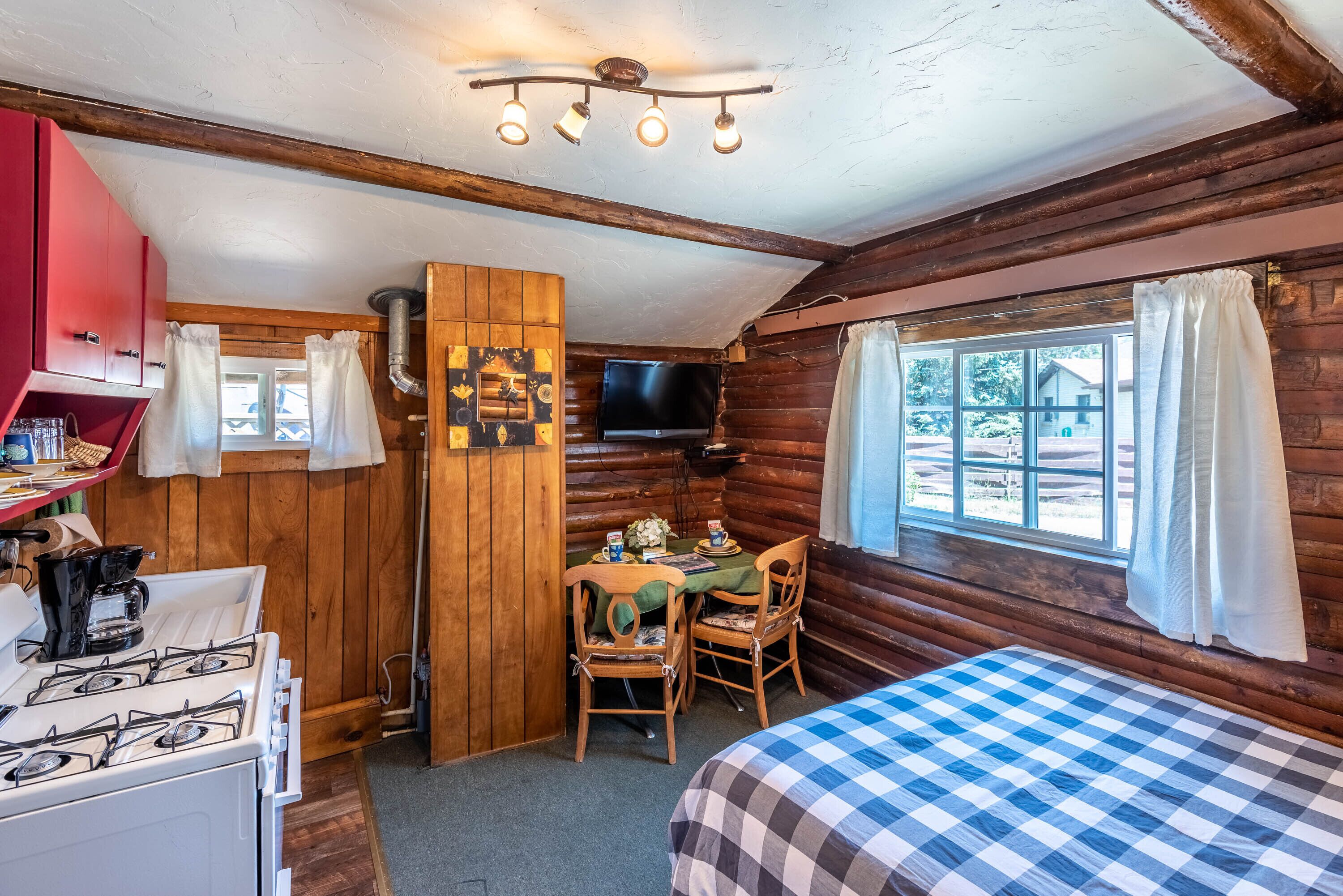 A cozy cabin-style room with a wooden interior, a dining table with chairs, a bed with a checkered blanket, a television, and a gas stove.