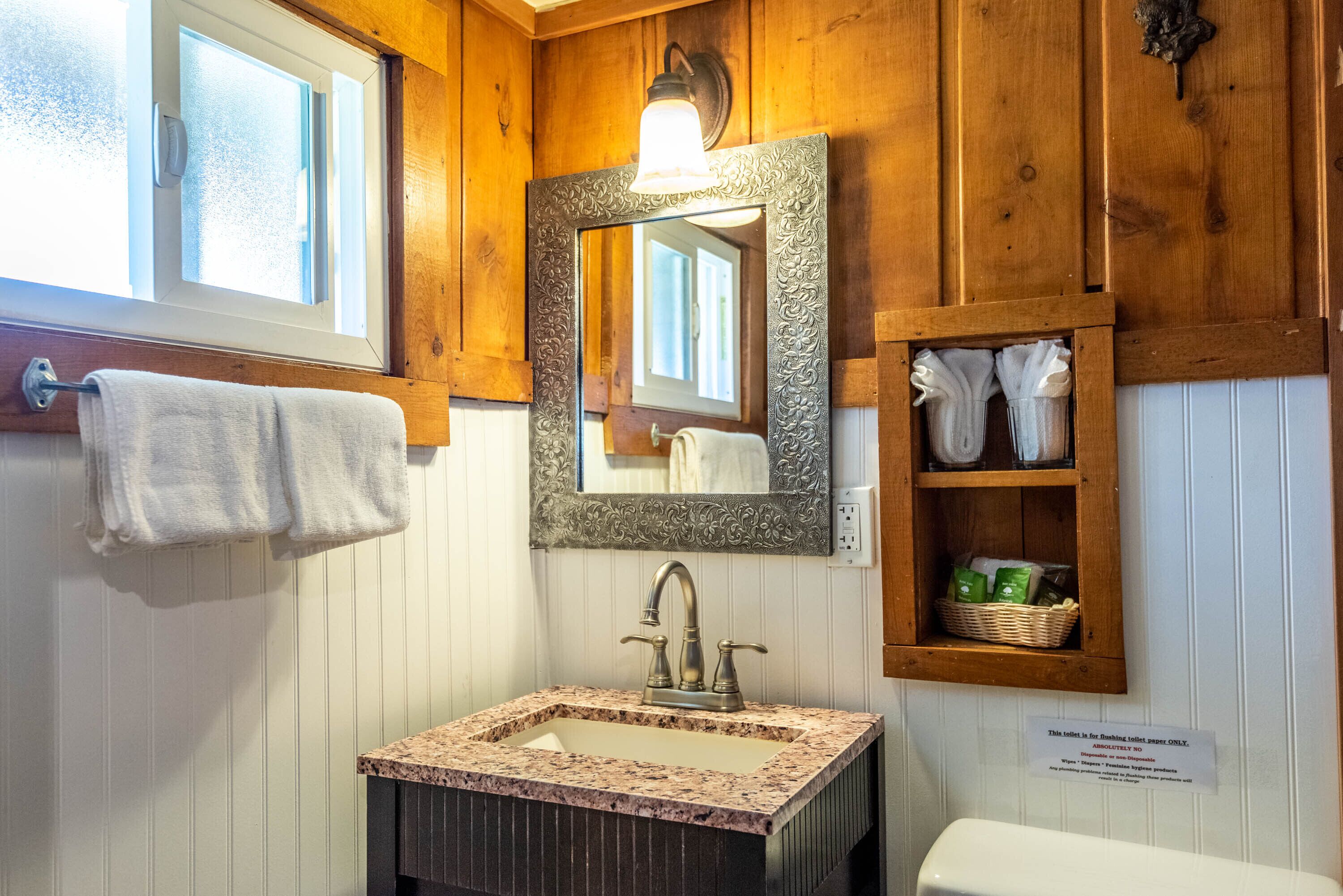 Une salle de bain avec un meuble-lavabo en bois, un évier surmonté d’un plan en marbre, un miroir avec un cadre orné et une étagère contenant des serviettes et des articles de toilette.